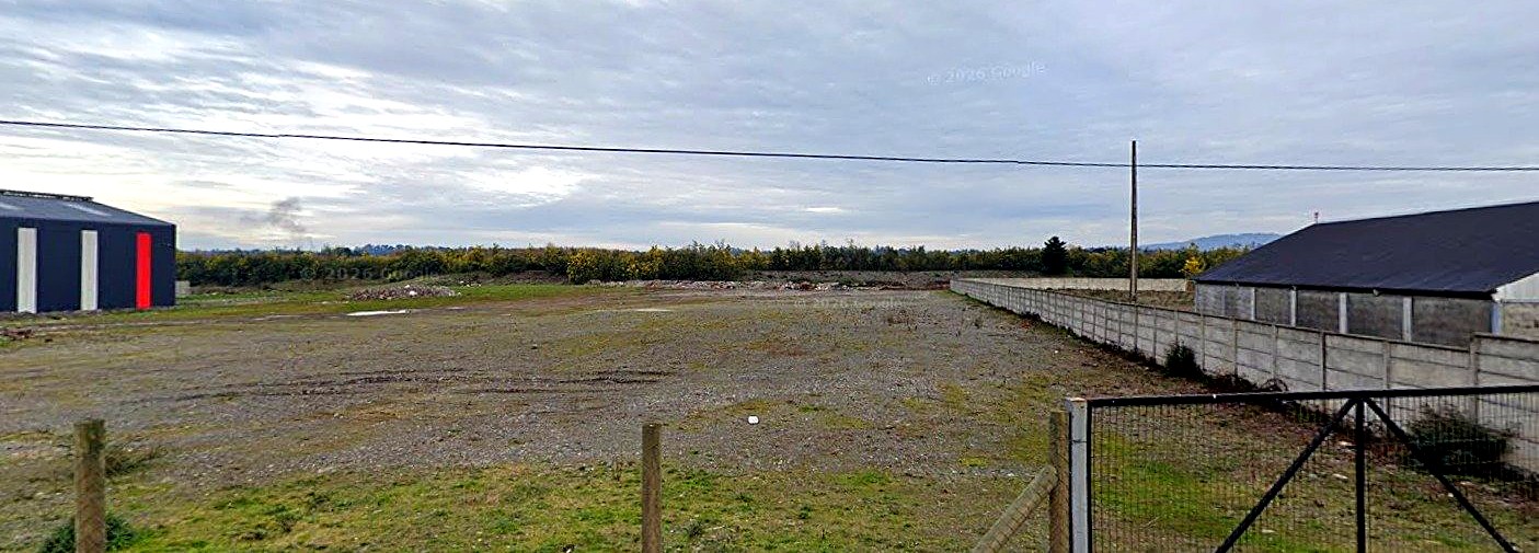 Vista real desde el camino: terreno visto desde el acceso público, con la bodega del predio vecino al fondo